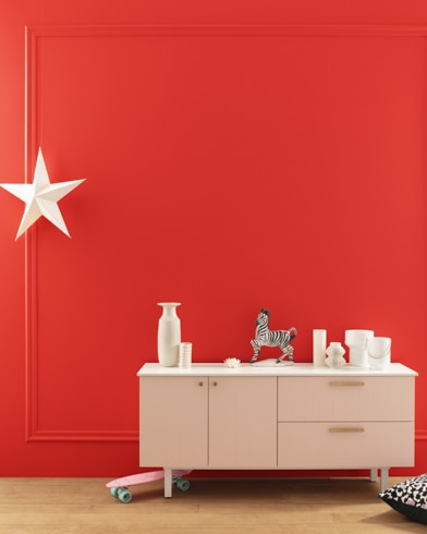 A modern white dresser with decor in front of a Vermilion-painted wall with square molding, and a dangling white star. 