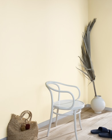 A palm frond, straw tote bag, black slide sandals and white metal chair lean against a wall painted Vanilla Ice Cream.