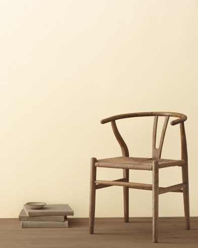 A modern wicker chair and a stack of books topped with a small bowl sit in front of a room painted Mannequin Cream.