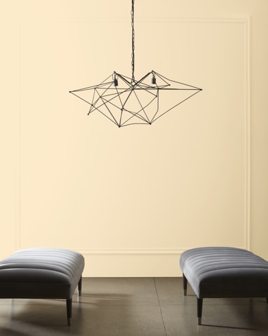 A modern, wiry chandelier hangs over two velvet ottomans in front of a wall painted Macadamia Nut.