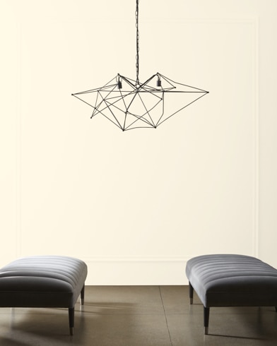 A modern, wiry chandelier hangs over two velvet ottomans in front of a wall painted Satin Shoes.