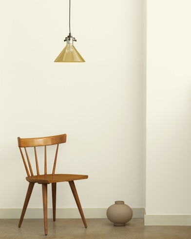 A single-bulb light hangs over a wooden chair and small ceramic pot in front of a wall painted Powder Sand.