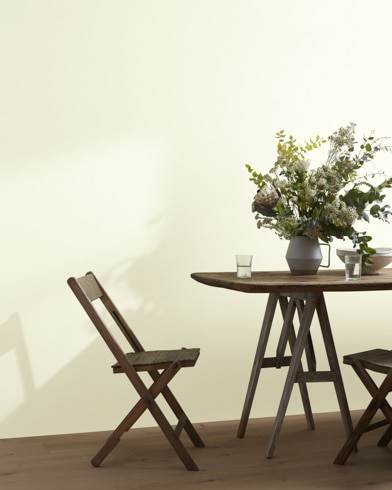 A wood folding chair sits next to a table topped with a greenery-filled vase in front of a Bavarian Cream-painted wall.