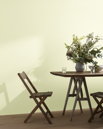 A wood folding chair sits next to a table topped with a greenery-filled vase in front of a Cream Silk-painted wall.