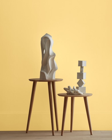 White abstract scupltures stand on wooden stools in front of a wall painted Crowne Hill Yellow.