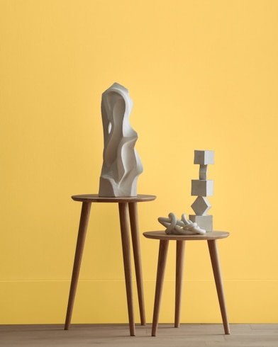 White abstract scupltures stand on wooden stools in front of a wall painted Golden Groves.