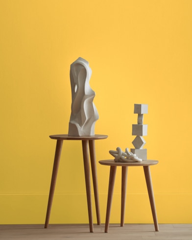 White abstract scupltures stand on wooden stools in front of a wall painted Imperial Yellow.