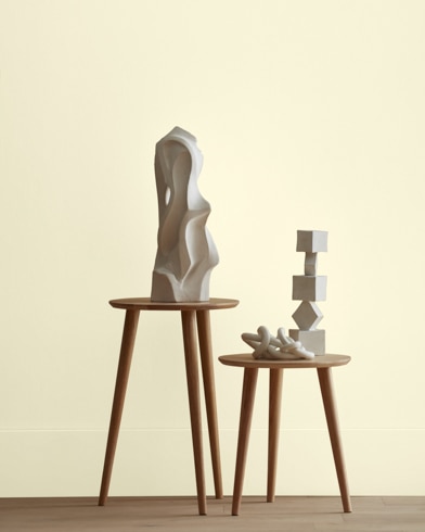 White abstract scupltures stand on wooden stools in front of a wall painted Yellow Freeze.