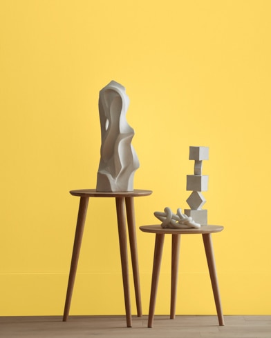 White abstract scupltures stand on wooden stools in front of a wall painted Yellow Raincoat.