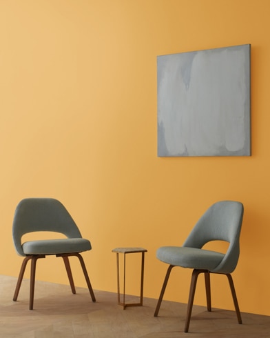 Two blue chairs and a table stand in front of a wall under an abstract painting hanging on a wall painted Glowing Umber.