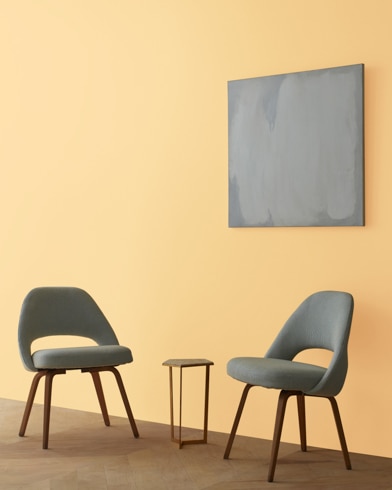 Two blue chairs and a table stand in front of a wall under an abstract painting hanging on a wall painted Moir Gold.
