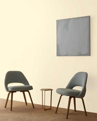 Two blue chairs and a table stand in front of a wall under an abstract painting hanging on a wall painted Ylang Ylang.