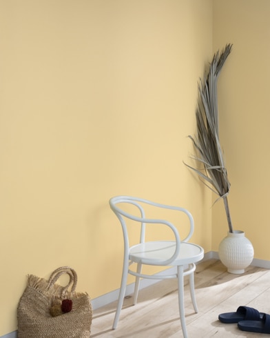 Una hoja de palma, un bolso de mano de paja, sandalias negras y una silla de metal blanca contra una pared pintada de color cebada.