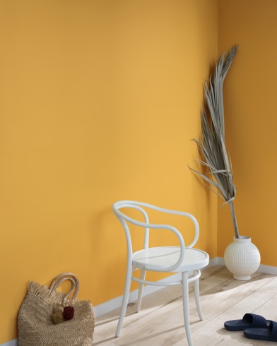 A palm frond, straw tote bag, black slide sandals and white metal chair lean against a wall painted Buttercup.