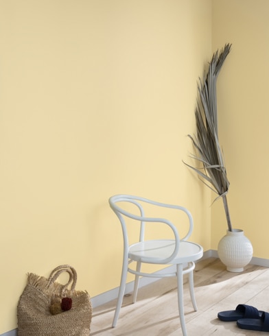 A palm frond, straw tote bag, black slide sandals and white metal chair lean against a wall painted Cornsilk.
