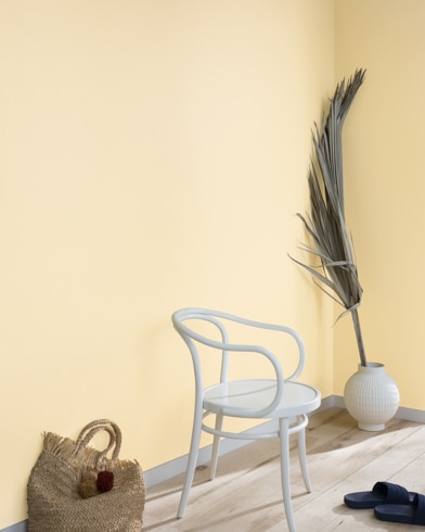 A palm frond, straw tote bag, black slide sandals and white metal chair lean against a wall painted Filtered Sunlight.