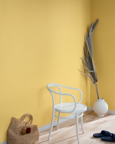 Una hoja de palma, un bolso de mano de paja, sandalias negras y una silla de metal blanca contra una pared pintada de color Hoja de Oro.