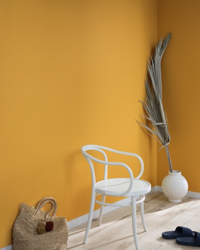 A palm frond, straw tote bag, black slide sandals and white metal chair lean against a wall painted Spicy Mustard.