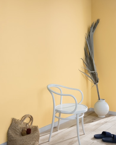 Una hoja de palma, un bolso de mano de paja, sandalias negras y una silla de metal blanca contra una pared pintada de color paja.