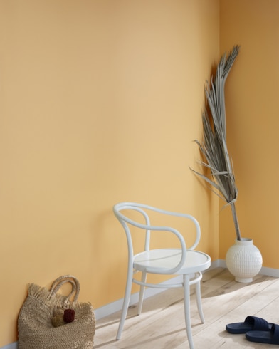 A palm frond, straw tote bag, black slide sandals and white metal chair lean against a wall painted Sweeney Yellow.