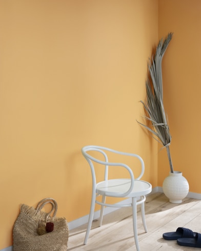 A palm frond, straw tote bag, black slide sandals and white metal chair lean against a wall painted Tavern Ochre.