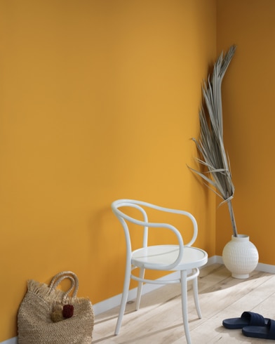 A palm frond, straw tote bag, black slide sandals and white metal chair lean against a wall painted Yellow Oxide.