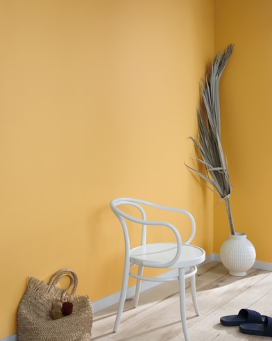 A palm frond, straw tote bag, black slide sandals and white metal chair lean against a wall painted York Harbor Yellow.