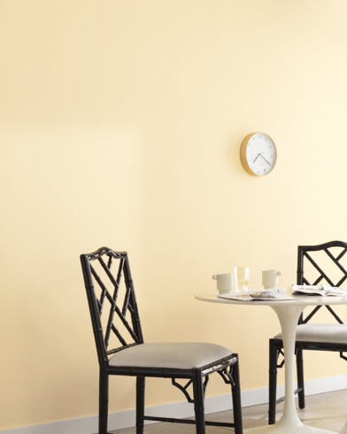 A clock hangs on a wall painted Aura in a room with a metal dining set and a potted plant on a wooden block.