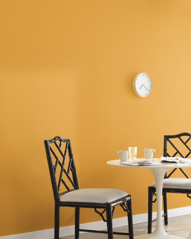 A clock hangs on a wall painted Mayan Gold in a room with a metal dining set and a potted plant on a wooden block.