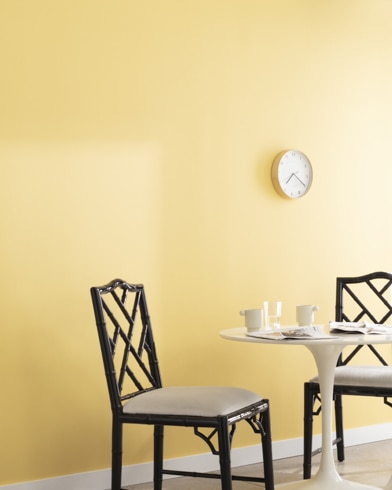 A clock hangs on a wall painted Sunny Days in a room with a metal dining set and a potted plant on a wooden block.