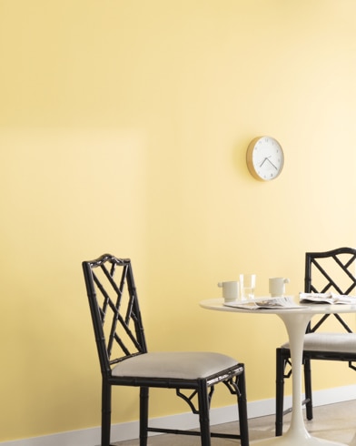 A clock hangs on a wall painted Traditional Yellow in a room with a metal dining set and a potted plant on a wooden block.