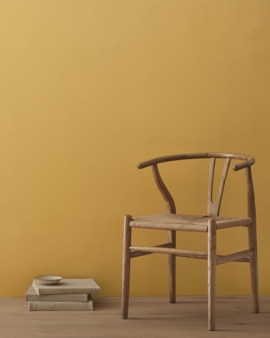A modern wicker chair and a stack of books topped with a small bowl sit in front of a room painted Autumn Gold.