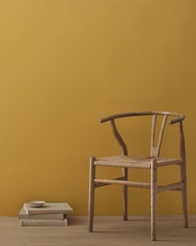 A modern wicker chair and a stack of books topped with a small bowl sit in front of a room painted Aztec Yellow.