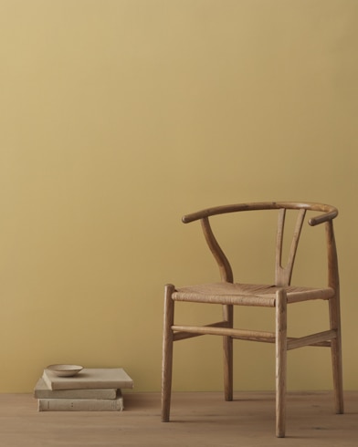 A modern wicker chair and a stack of books topped with a small bowl sit in front of a room painted Blair Gold.