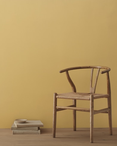 A modern wicker chair and a stack of books topped with a small bowl sit in front of a room painted Citrine.