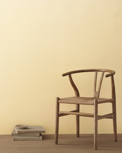 A modern wicker chair and a stack of books topped with a small bowl sit in front of a room painted Golden Straw.