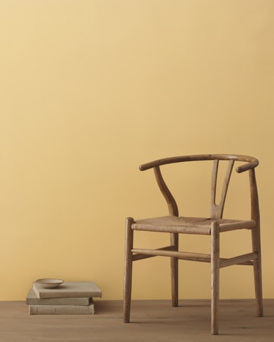 A modern wicker chair and a stack of books topped with a small bowl sit in front of a room painted Golden Tan.