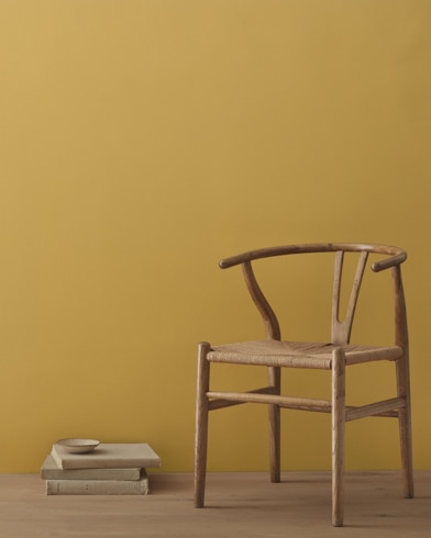 A modern wicker chair and a stack of books topped with a small bowl sit in front of a room painted Palace Ochre.