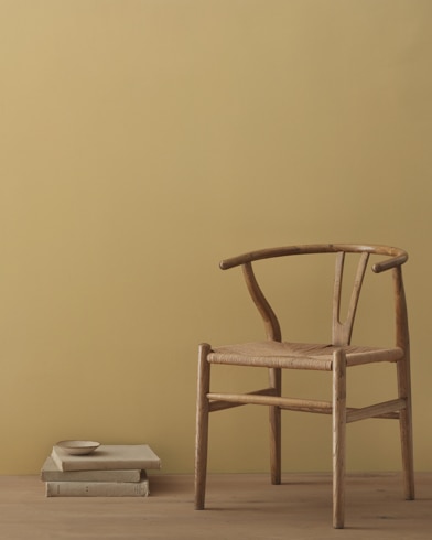 A modern wicker chair and a stack of books topped with a small bowl sit in front of a room painted Scrivener Gold.