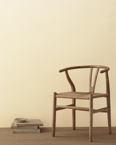 A modern wicker chair and a stack of books topped with a small bowl sit in front of a room painted Simply Irresistible.