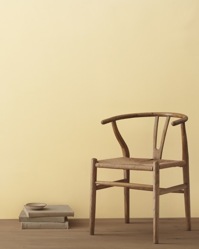 Una moderna silla de mimbre y una pila de libros con un pequeño bol apoyado delante de una pared pintada de color Cosecha del Verano.
