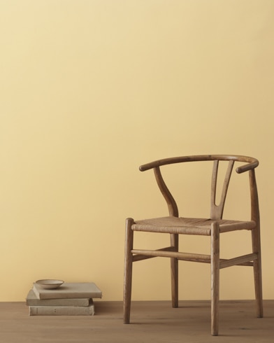 A modern wicker chair and a stack of books topped with a small bowl sit in front of a room painted Vellum.
