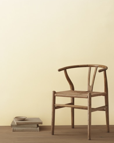 Una moderna silla de mimbre y una pila de libros con un pequeño bol apoyado delante de una pared pintada de color Crema de Woodmont.