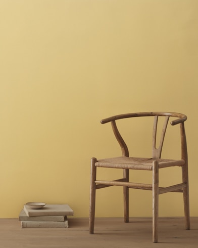 A modern wicker chair and a stack of books topped with a small bowl sit in front of a room painted Wythe Gold.