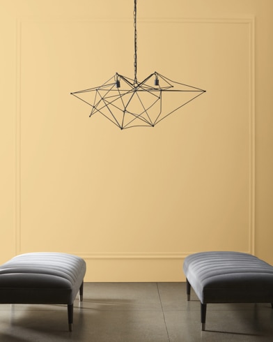 A modern, wiry chandelier hangs over two velvet ottomans in front of a wall painted Dijon.