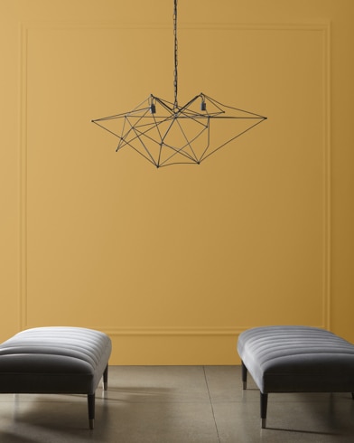A modern, wiry chandelier hangs over two velvet ottomans in front of a wall painted French Horn.