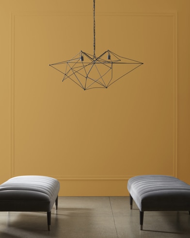 A modern, wiry chandelier hangs over two velvet ottomans in front of a wall painted Goldenhurst.