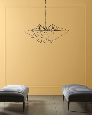 A modern, wiry chandelier hangs over two velvet ottomans in front of a wall painted Hathaway Gold.