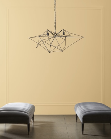A modern, wiry chandelier hangs over two velvet ottomans in front of a wall painted Shortbread.