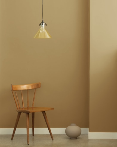 A single-bulb light hangs over a wooden chair and small ceramic pot in front of a wall painted Everard Gold.